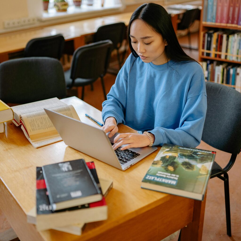 Current image: Asian woman typing on a laptop surrounded by books in a library setting.